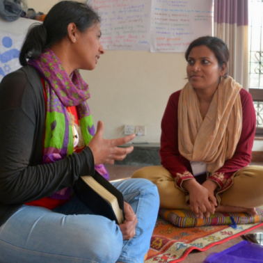 Two South Asian women sit cross-legged in a small room to speak