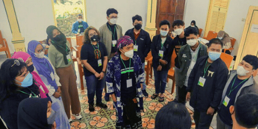 Group of people, of mixed ages and genders, some with headscarves, stand in a circle wearing masks as one person in the center speaks
